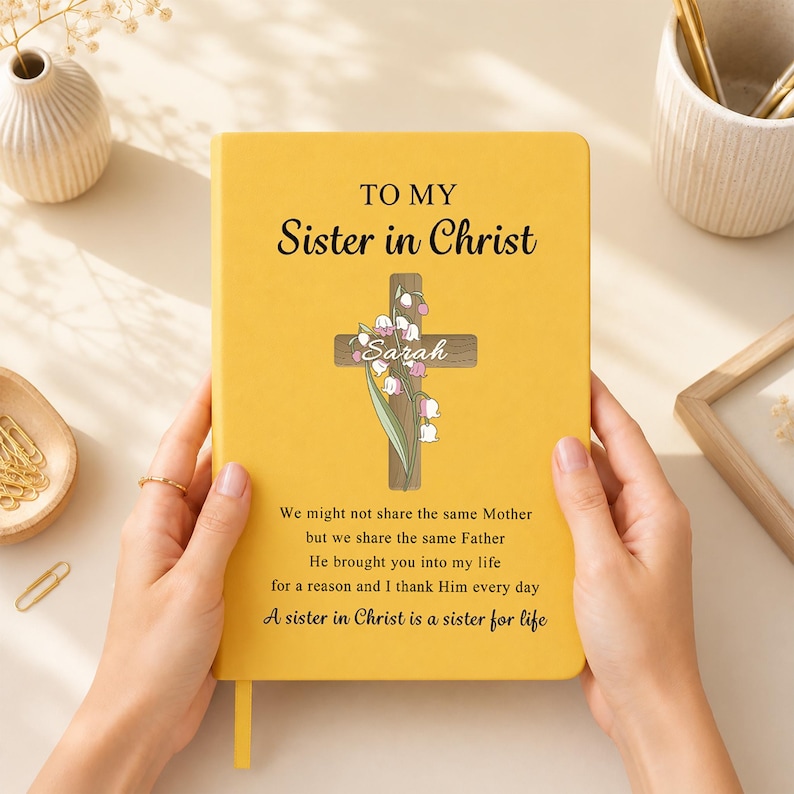 May include: A yellow hardcover journal featuring the text TO MY Sister in Christ, a wooden cross graphic with lily of the valley flowers, the name Sarah, and a printed poem about sisterhood. The journal has a small yellow ribbon bookmark.