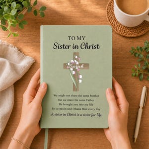 May include: A sage green journal featuring the text TO MY Sister in Christ, a wooden cross graphic with lily of the valley flowers, the name Sarah, and a poem that reads We might not share the same Mother but we share the same Father.