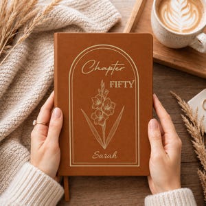 May include: A rectangular brown notebook featuring a gold-colored line art illustration of a flower centered within an arched frame. The text "Chapter FIFTY" is printed above the flower, and the name "Sarah" is printed below in cursive script.