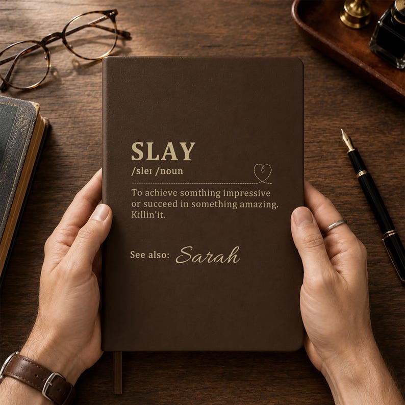 May include: A dark brown notebook featuring gold text that reads SLAY /sleɪ /noun To achieve somthing impressive or succeed in something amazing. Killin'it. See also: Sarah. The cover includes a small stitched heart graphic above the definition.