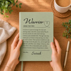 Peut inclure: Un carnet vert sauge pr&eacute;sentant la d&eacute;finition du mot Warrior en texte noir, incluant une ic&ocirc;ne de c&oelig;ur et le nom Sarah en bas. La couverture affiche un paragraphe d&eacute;crivant un guerrier comme un esprit de courage et de r&eacute;silience.
