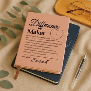 May include: A peach-colored notebook with the text Difference Maker, a definition, and the name Sarah printed in black. A dark blue notebook lies underneath it, accompanied by a clear fountain pen and a pair of round glasses nearby.