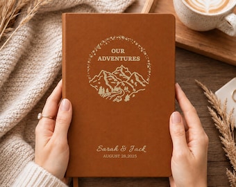 May include: A brown leather-bound journal featuring a gold-colored circular graphic of mountains, trees, and stars. The text reads "OUR ADVENTURES" above the mountain illustration, with "Sarah & Jack" and "AUGUST 28, 2025" printed in gold below.