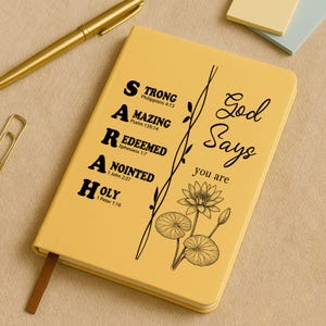 Peut inclure: Un cahier jaune avec les mots "SARAH" et "Dieu dit que tu es" imprimés sur la couverture. Le mot "SARAH" est épelé verticalement avec des mots descriptifs et des versets bibliques. Un stylo et un trombone dorés sont visibles.