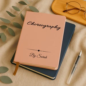 May include: A peach-colored notebook with the word "Choreography" in script, and "By Sarah" below. A dark blue notebook is beneath it. A gold bookmark and a pen are visible. The notebooks are on a beige surface.