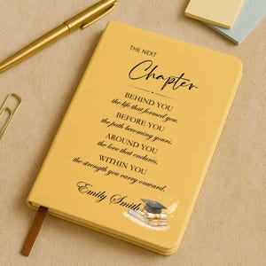 May include: Mustard yellow hardcover journal with the text "The Next Chapter" and inspirational quotes. Features a graduation cap and scroll illustration. Includes a brown ribbon bookmark and the name "Emily Smith". A gold pen and paperclip are in the background.