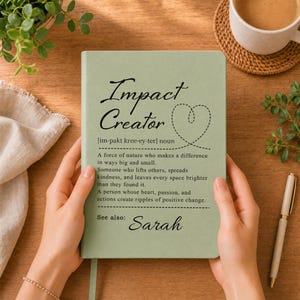 May include: A sage green notebook featuring the black text Impact Creator, a phonetic pronunciation, a definition of the term, a dashed heart outline, and the name Sarah printed at the bottom. The cover has a smooth, matte finish.
