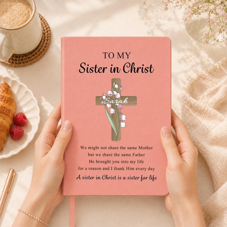 May include: A coral-colored notebook featuring the text "TO MY Sister in Christ" above a wooden cross illustration adorned with lily of the valley flowers and the name "Sarah". Below, the text reads "We might not share the same Mother but we share the same Father. He brought you into my life for a reason and I thank Him every day. A sister in Christ is a sister for life."