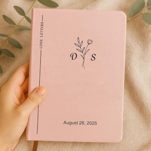 May include: A pink journal with the text "LOVE LETTERS" on the spine. The cover features a floral design with the initials "D" and "S", and the date "August 28, 2025". The journal is held by a hand.