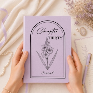 May include: A light purple hardback journal featuring a black line art illustration of a gladiolus flower inside an arched frame. The text "Chapter THIRTY" is printed above the flower, and the name "Sarah" is printed below in black script.