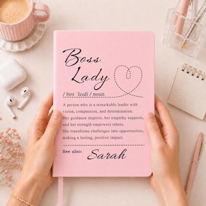 May include: A light pink notebook featuring the black text Boss Lady, a heart outline, and a dictionary-style definition describing a leader with vision and determination, followed by the name Sarah at the bottom of the front cover.