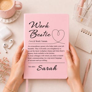 Könnte beinhalten: Ein hellrosa Notizbuch mit dem schwarzen Aufdruck Work Bestie, einer Herz-Kontur, einer phonetischen Aussprache, einer humorvollen Definition eines Arbeitskollegen und dem Namen Sarah unten, gehalten in vertikaler Ausrichtung.