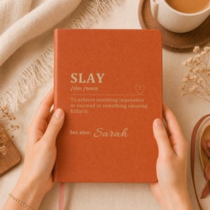 May include: A rust-colored notebook with a matte finish featuring the white printed definition of the word SLAY, including the phonetic spelling, a heart icon, and the text See also: Sarah, centered on the front cover in a clean, sans-serif font.