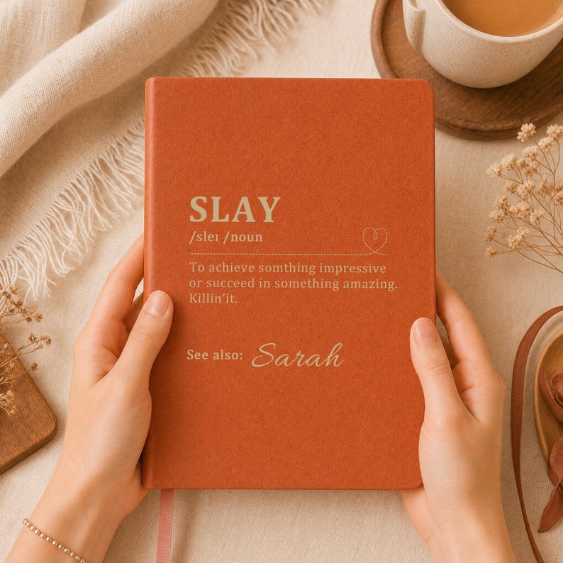May include: A rust-colored notebook with a matte finish featuring the white printed definition of the word SLAY, including the phonetic spelling, a heart icon, and the text See also: Sarah, centered on the front cover in a clean, sans-serif font.