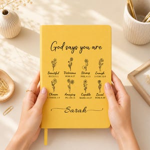 May include: A yellow notebook featuring the black text God says you are, followed by eight floral illustrations paired with words like Beautiful, Victorious, Strong, Enough, Chosen, Amazing, Capable, and Loved, with the name Sarah printed below.