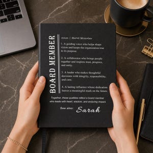 Op de afbeelding: Een zwart notitieboek met witte tekst met een woordenboekdefinitie van de term BOARD MEMBER, met vier beschrijvende punten over leiderschap en invloed, gevolgd door de naam Sarah onder het kopje See also.