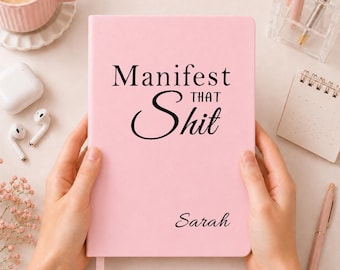 May include: A light pink notebook with black text that reads Manifest THAT Shit and the name Sarah printed in a cursive font. The notebook has a smooth, matte cover and a vertical ribbon bookmark extending from the bottom edge of the spine.