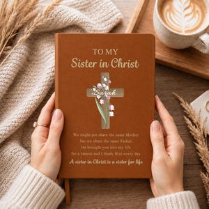 May include: A brown notebook featuring the text TO MY SISTER IN CHRIST, a wooden cross graphic with lily of the valley flowers, the name Sarah, and the quote We might not share the same Mother but we share the same Father. A sister in Christ is a sister for life.