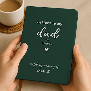 Pode incluir: Um diário verde escuro com o texto "Letters to my dad in Heaven" e "in loving memory of Sarah" em escrita branca. Um símbolo de coração branco também está presente. O diário é segurado por uma pessoa.