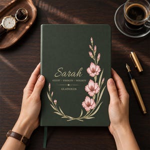 May include: A dark green notebook featuring a floral illustration of pink gladiolus flowers on the right side. The text reads Sarah, August, Strength, Integrity, and Gladiolus. The cover has a smooth matte finish and a thin green ribbon bookmark.