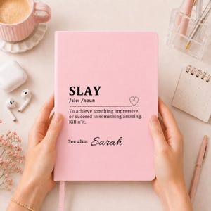 May include: A light pink notebook featuring the black printed text SLAY /sleɪ /noun followed by the definition To achieve somthing impressive or succeed in something amazing. Killin'it. and the text See also: Sarah with a small heart icon.
