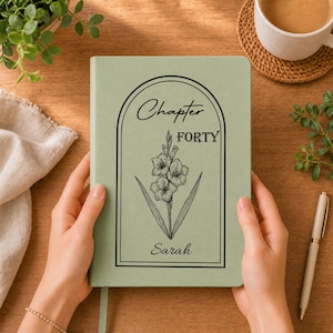 May include: A sage green hardcover journal featuring a black line art illustration of a gladiolus flower centered within an arched frame. The text "Chapter FORTY" appears above the flower, and the name "Sarah" is printed in script below it.