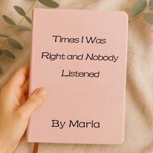 May include: A pale pink notebook featuring the text "Times I Was Right and Nobody Listened" and the author's name "By Maria" in black. The journal is held in a hand, set against a beige backdrop.