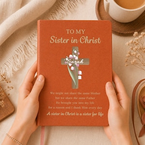 May include: A burnt orange notebook featuring the text TO MY Sister in Christ, a wooden cross illustration with lily of the valley flowers, the name Sarah, and the poem: We might not share the same Mother but we share the same Father.