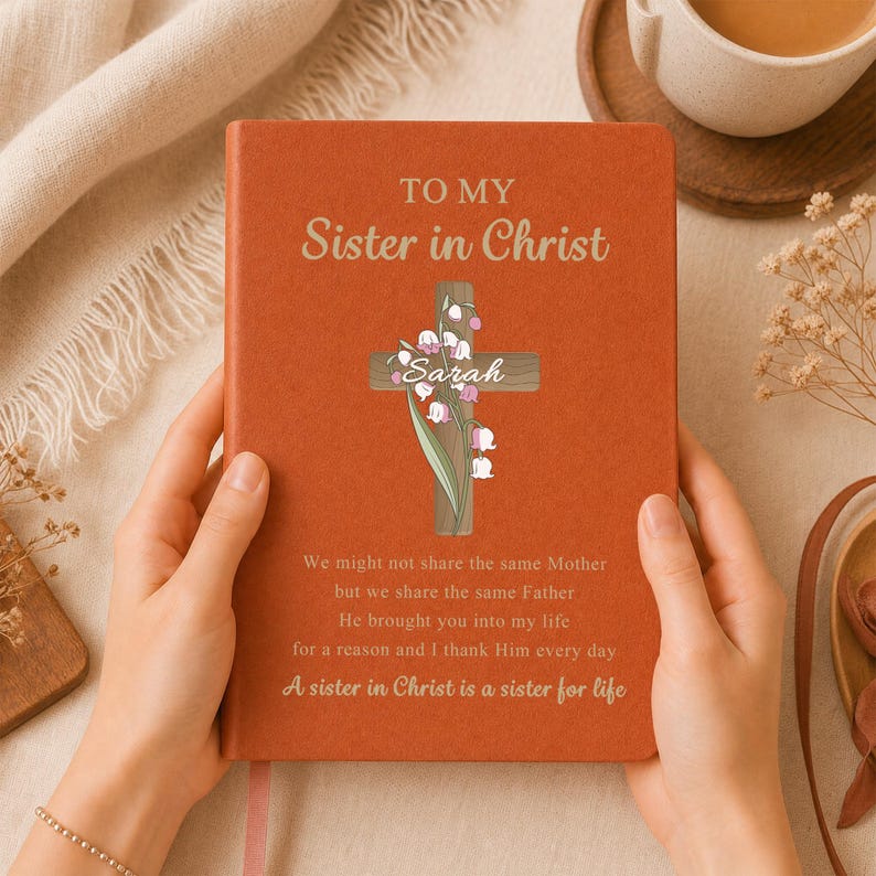 May include: A burnt orange notebook featuring the text TO MY Sister in Christ, a wooden cross illustration with lily of the valley flowers, the name Sarah, and the poem: We might not share the same Mother but we share the same Father.