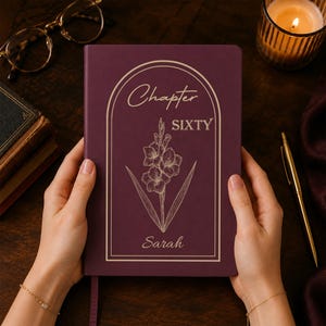 May include: A deep purple hardback journal featuring a gold-coloured arched frame, a line-art illustration of a gladiolus flower, and the text "Chapter SIXTY" above the name "Sarah" printed in a gold-coloured script font on the front cover.