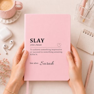 Puede incluir: Un cuaderno rosa claro con el texto impreso en negro SLAY /sleɪ /noun, seguido de la definición: To achieve something impressive or succeed in something amazing. Killin'it. Debajo del texto, se lee: See also: Sarah.
