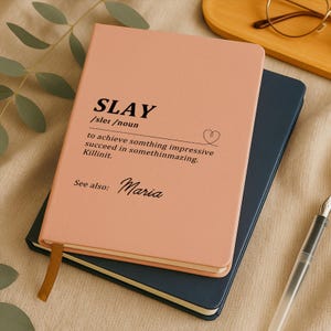 Könnte beinhalten: Ein rosa Notizbuch mit dem Wort „SLAY“ auf dem Cover, zusammen mit einer Definition und dem Namen „Maria“. Das Notizbuch liegt auf einem marineblauen Notizbuch. Ein goldener Lesezeichen ist sichtbar.