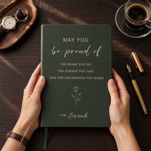 May include: A dark green notebook with gold foil text that reads: MAY YOU be proud of THE WORK YOU DO THE PERSON YOU ARE AND THE DIFFERENCE YOU MAKE, followed by a line drawing of a rose and the name Sarah at the bottom of the cover.