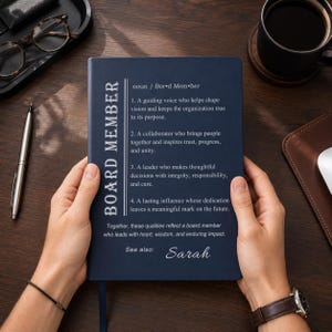 May include: A dark blue notebook featuring the white printed definition of a board member, listing four descriptive points about leadership and collaboration, followed by the text 'See also: Sarah' in a white script font on the front cover.