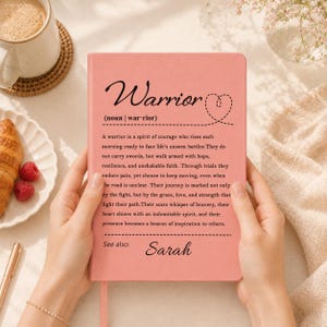 Op de afbeelding: Een rechthoekig roze notitieboek met de gedrukte tekst Warrior (noun | war-rior) gevolgd door een definitie van een krijger en de tekst See also: Sarah. De kaft bevat een gestippelde hartomtrek en zwarte tekst op een roze achtergrond.