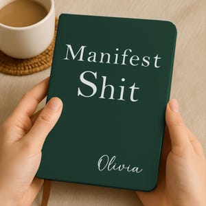 Puede incluir: Un cuaderno verde oscuro con las palabras "Manifest Shit" en blanco y el nombre "Olivia" en letra cursiva. El cuaderno está en manos de alguien, con una taza de café en el fondo.