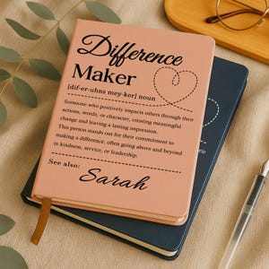 May include: A pink notebook with the words "Difference Maker" and a definition printed on the cover. The notebook is stacked on top of a dark blue notebook. A gold ribbon bookmark is visible.
