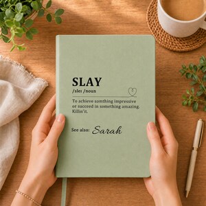 May include: A sage green hardcover notebook featuring black printed text that defines the word SLAY as achieving something impressive or succeeding in something amazing, with the additional text See also: Sarah and a small heart icon.