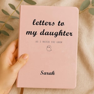 Könnte beinhalten: Ein rosa Notizbuch mit dem Titel "letters to my daughter" und dem Untertitel "as I watch you grow". Das Cover zeigt eine Geisterillustration und den Namen "Sarah". Das Notizbuch wird von einer Hand gehalten.