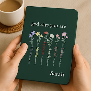 May include: A dark green journal held by hands, featuring the text "god says you are" above floral illustrations and words like "Chosen", "Strong", and "Unique". The name "Sarah" is printed at the bottom. A cup of coffee sits in the background.