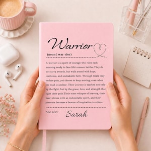 Peut inclure: Un carnet rose p&acirc;le avec la d&eacute;finition du mot Warrior imprim&eacute;e en noir, incluant un symbole de c&oelig;ur et le pr&eacute;nom Sarah en bas. Le texte d&eacute;crit un esprit de courage, de r&eacute;silience et d'espoir, le tout sur une couverture rose unie.
