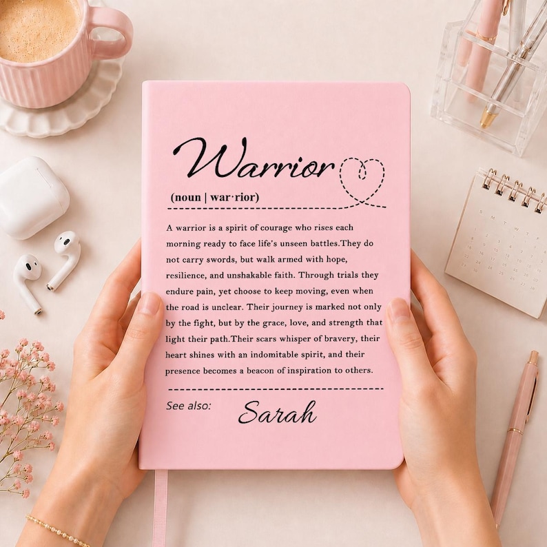 Peut inclure: Un carnet rose p&acirc;le avec la d&eacute;finition du mot Warrior imprim&eacute;e en noir, incluant un symbole de c&oelig;ur et le pr&eacute;nom Sarah en bas. Le texte d&eacute;crit un esprit de courage, de r&eacute;silience et d'espoir, le tout sur une couverture rose unie.