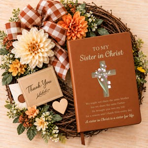Op de afbeelding: Een bruin lederen dagboek met de tekst TO MY Sister in Christ, een kruisafbeelding met de naam Sarah en een gedicht. Het ligt op een krans met oranje en crèmekleurige bloemen, een bruin geruit lint, een Thank You kaartje en een houten hart.
