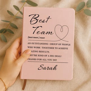 May include: A light pink notebook with black text that reads Best Team, followed by a dictionary-style definition, a dashed heart outline, and the name Sarah printed at the bottom. The cover features a smooth, matte finish and a ribbon bookmark.