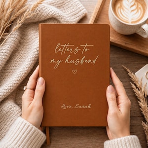 Op de afbeelding: Een rechthoekig notitieboek met een gladde, effen bruine kaft. De voorzijde is voorzien van goudkleurige cursieve tekst met "letters to my husband" boven een klein hartje, met de tekst "Love, Sarah" in goud gedrukt bij de onderrand.