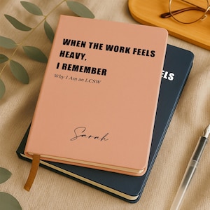 May include: A pink notebook with the text "WHEN THE WORK FEELS HEAVY, I REMEMBER Why I Am an LCSW" and the name "Sarah" in script. A dark blue notebook is beneath it. A gold ribbon bookmark is visible.