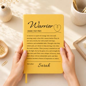 Peut inclure: Un carnet jaune avec du texte noir indiquant Warrior (noun | war-rior) suivi d'un paragraphe d&eacute;finissant un guerrier comme un esprit de courage, et le texte See also: Sarah en bas. La couverture pr&eacute;sente une ic&ocirc;ne de c&oelig;ur en pointill&eacute;s.