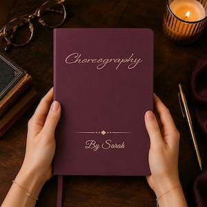 May include: A deep purple hardcover notebook featuring the gold-colored text Choreography By Sarah on the front cover. The cover includes a decorative horizontal line with a small diamond shape in the center, positioned between the two lines of text.