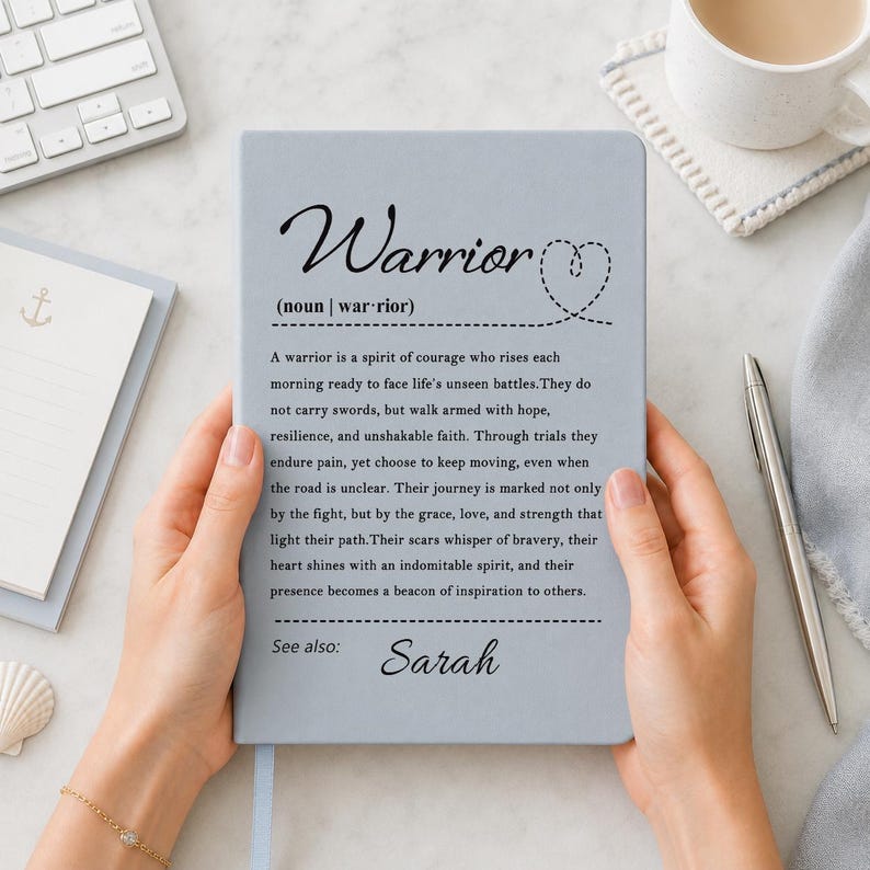 Peut inclure: Un carnet bleu clair pr&eacute;sentant la d&eacute;finition du mot Warrior en texte noir, incluant une ic&ocirc;ne de c&oelig;ur en pointill&eacute;s et le nom Sarah imprim&eacute; en bas. Le texte d&eacute;crit un guerrier comme un esprit de courage, de r&eacute;silience et de foi.