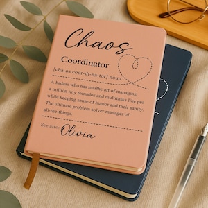 May include: A pink notebook with the word "Chaos" in cursive and the definition of "Coordinator". The notebook is stacked on top of a dark blue notebook. A gold ribbon bookmark is visible.